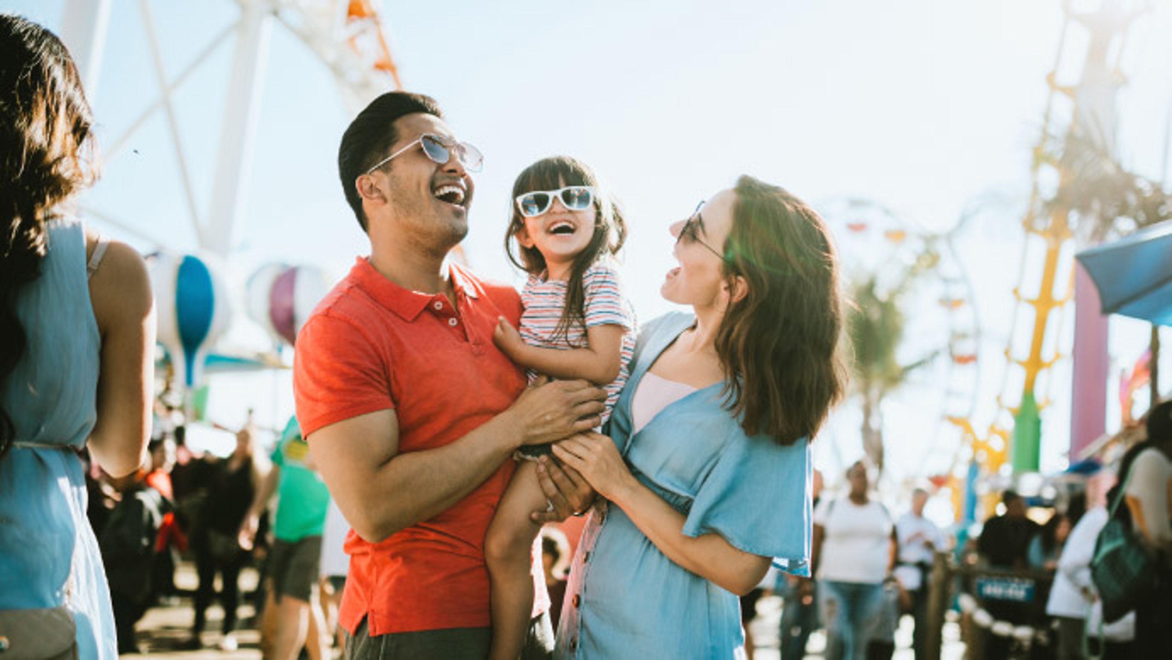 Man and a woman holding a young child in a lively outdoor setting, likely an amusement park or fairground.