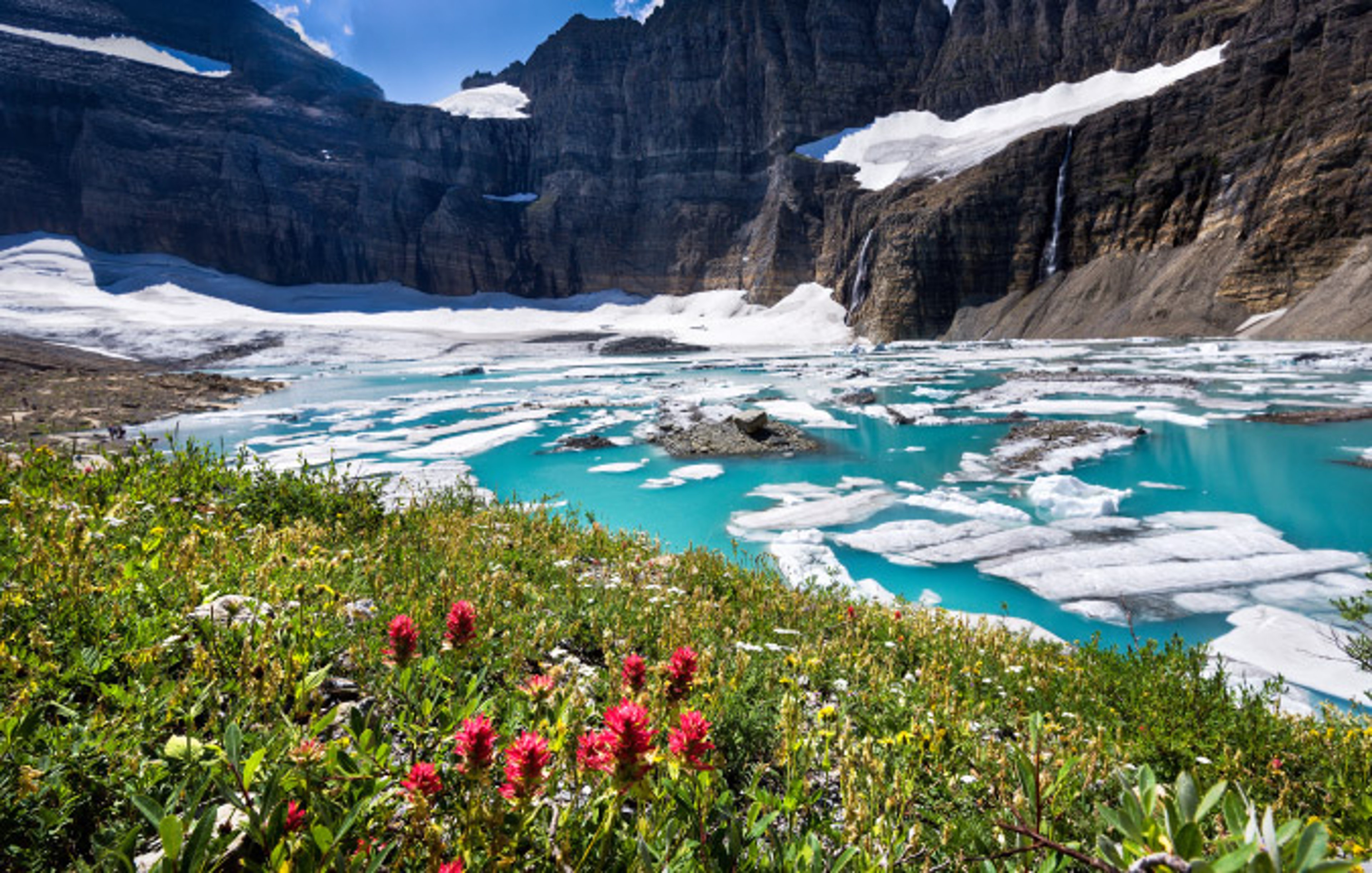 Turquois blue waters dotted white patches of ice at Grinnell Glacier in Glacier National Park, Montana.