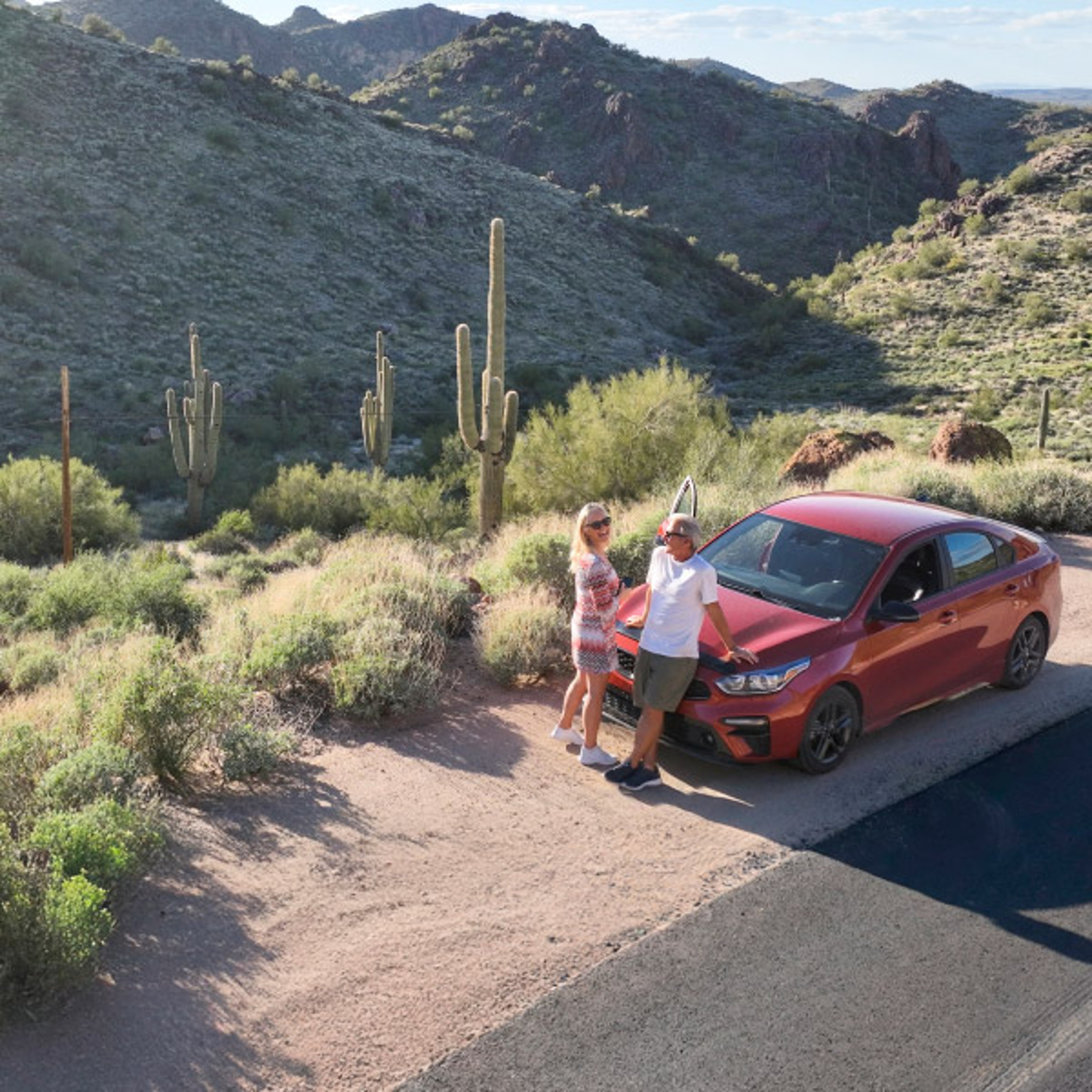 Couple leaning against the front of a red car while parked alongside the highway with dessert landscape in the background.