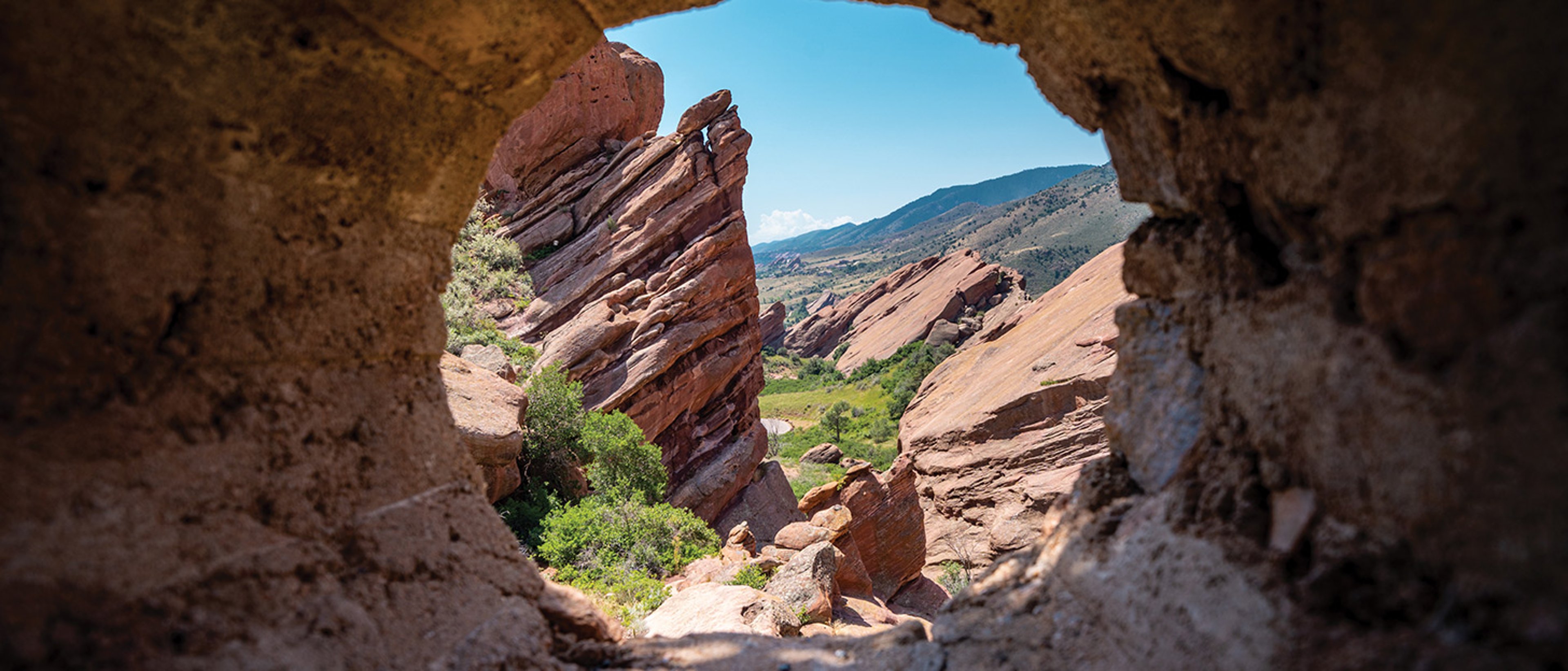 Photo of Red Rocks in Colorado that seems to be taken through a natural or carved rock opening, framing the view of the dramatic rock structures and the valley below