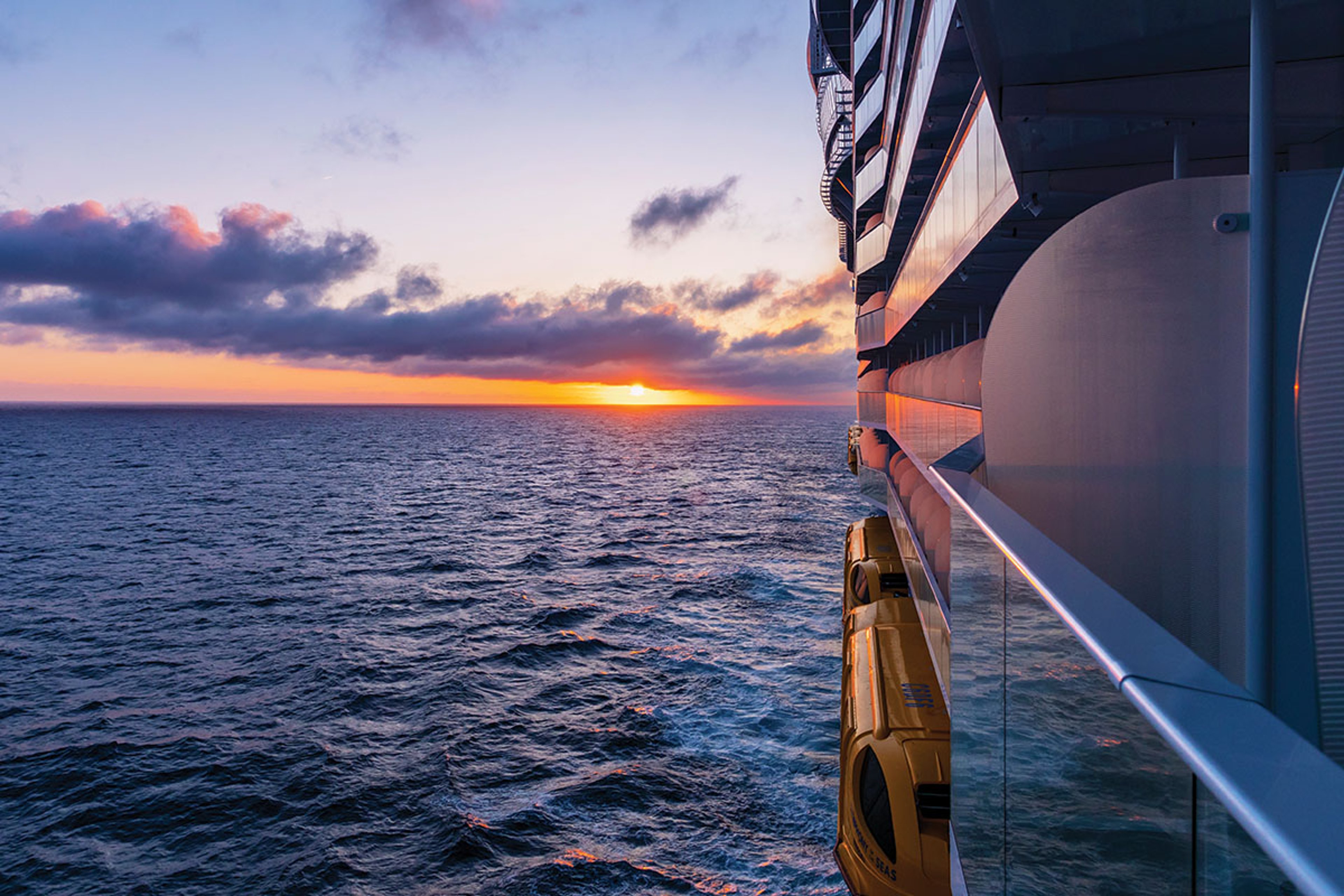 A vibrant orange and purple sunset over a dark blue, wavy ocean, viewed from the private balcony of a large Royal Caribbean cruise ship, with a yellow lifeboat visible on the right.