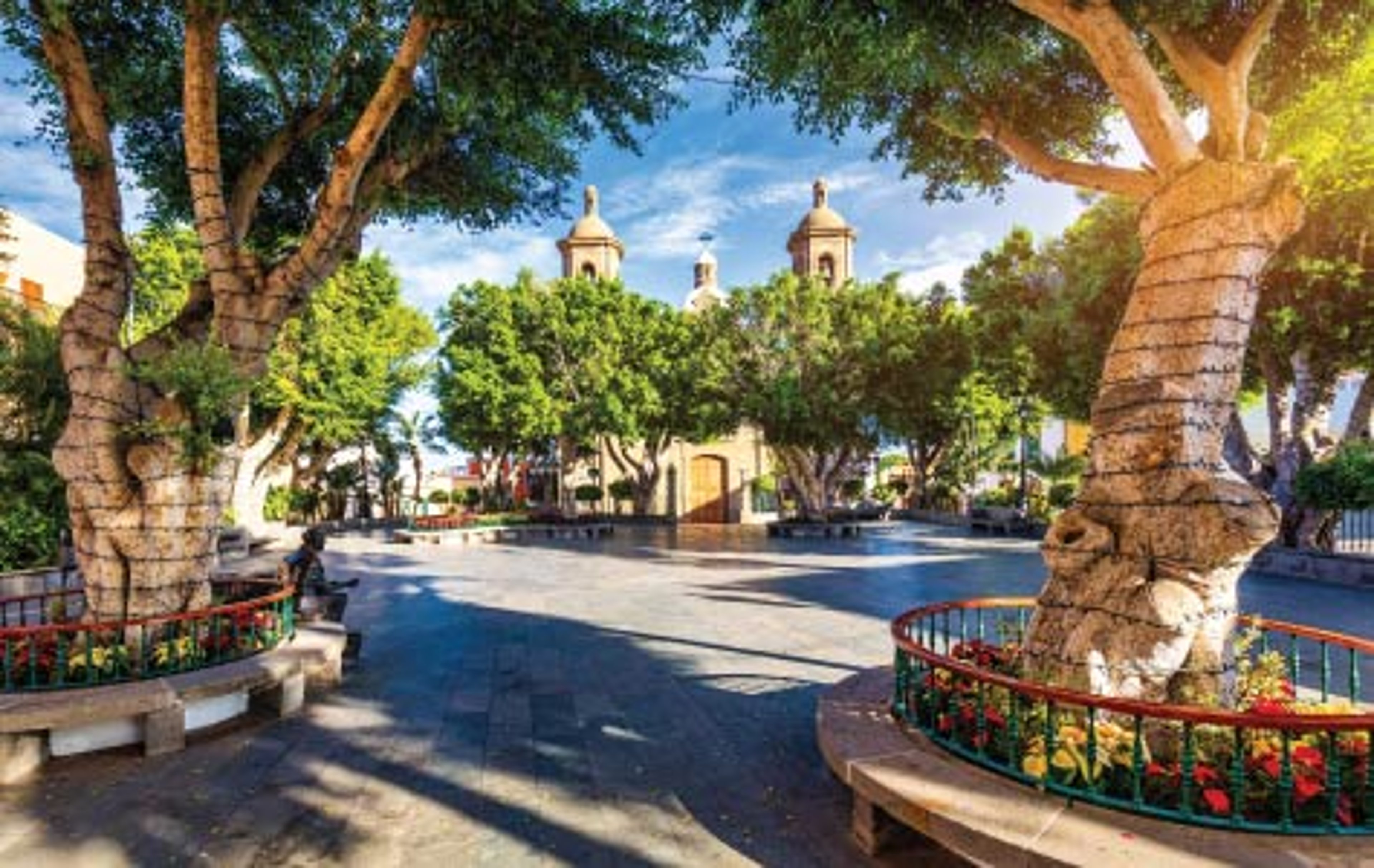 Sunlit town plaza in northern Spain with large sculptural trees in circular benches, colorful flower beds, and a historic church with twin bell towers framed by greenery under a blue sky.