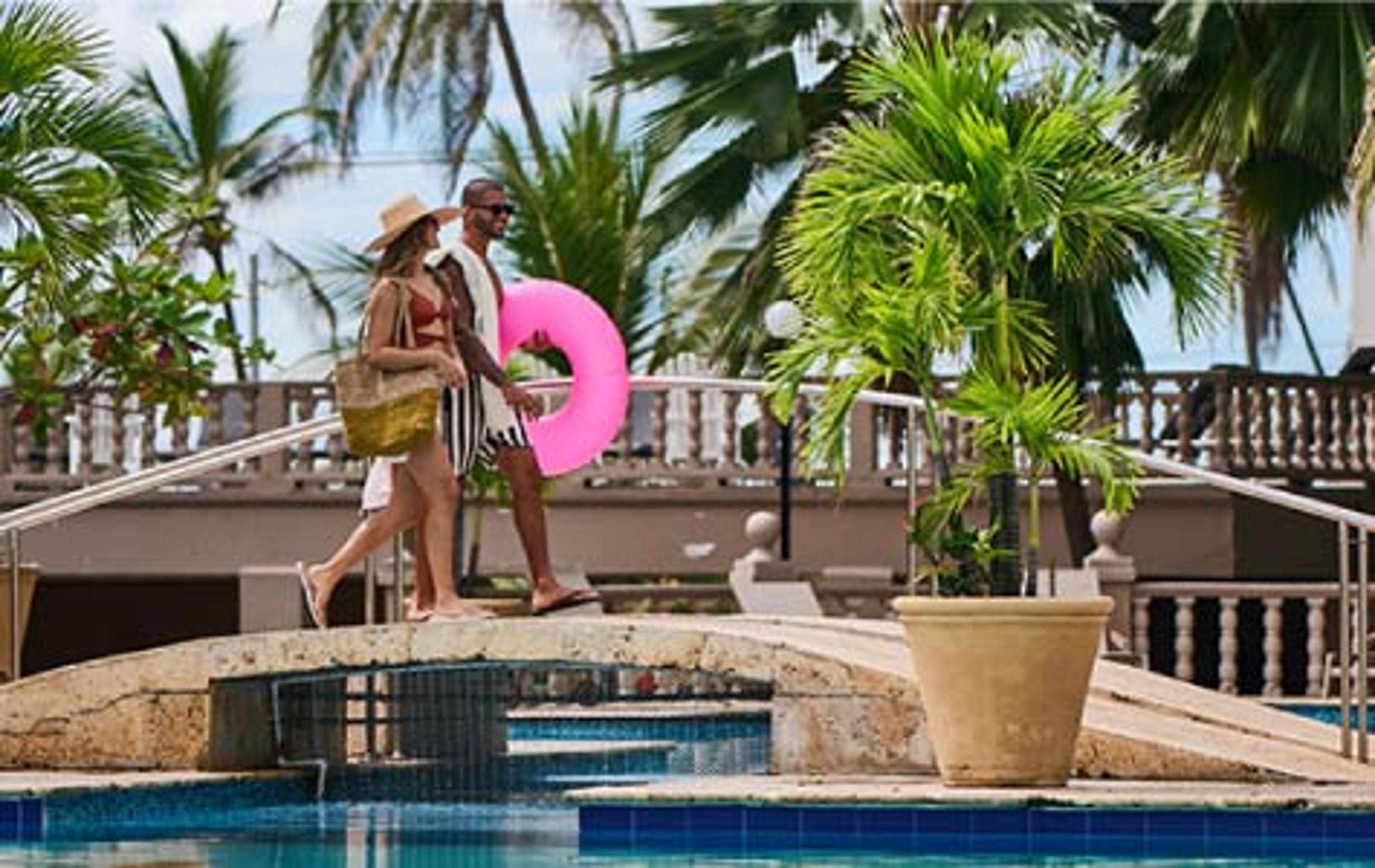 Couple crossing a bridge over a tropical resort pool, carrying a beach bag and pink inflatable ring with palm trees and ocean in the background.