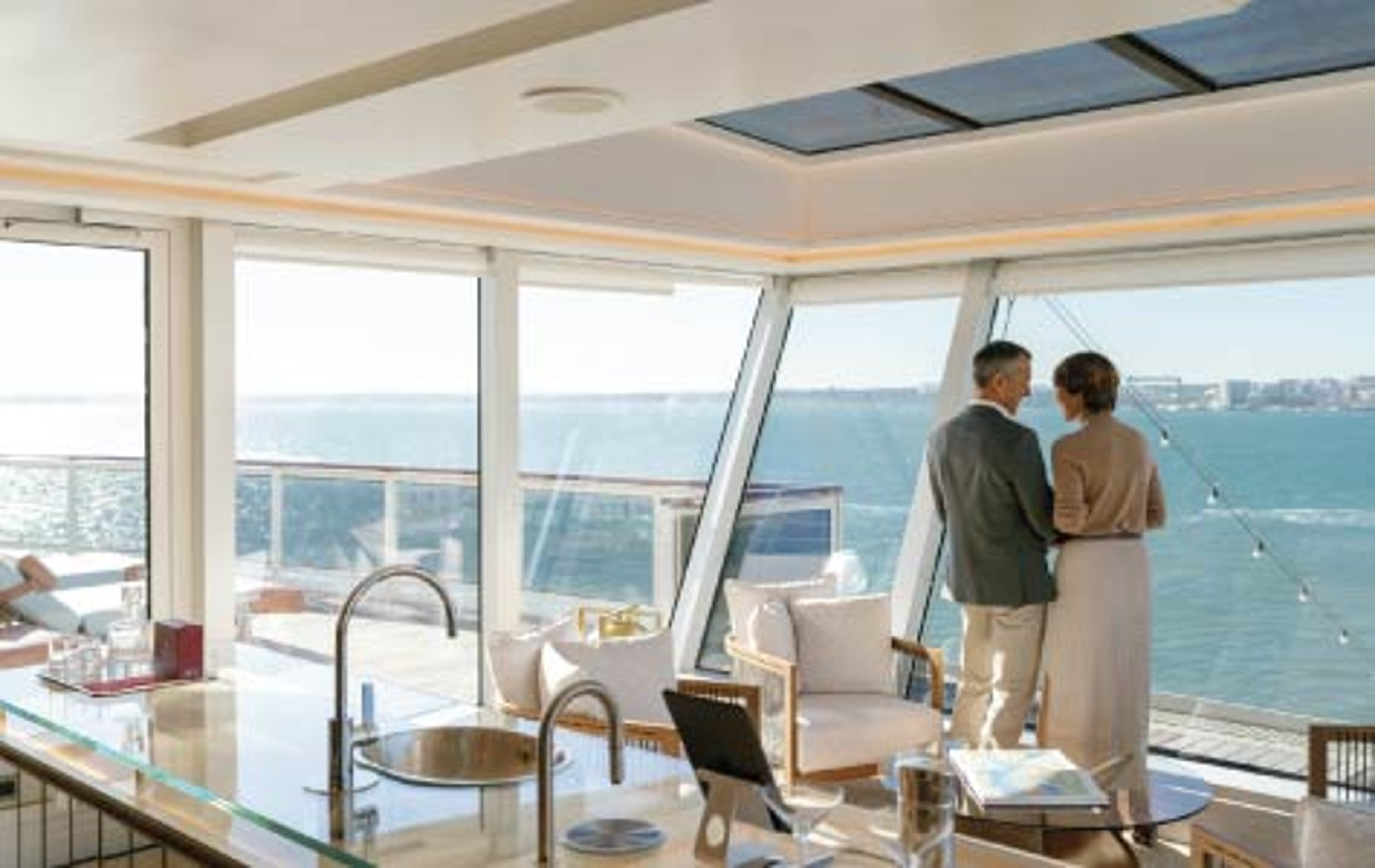 Couple standing inside a luxury cruise suite with floor-to-ceiling windows overlooking the ocean.