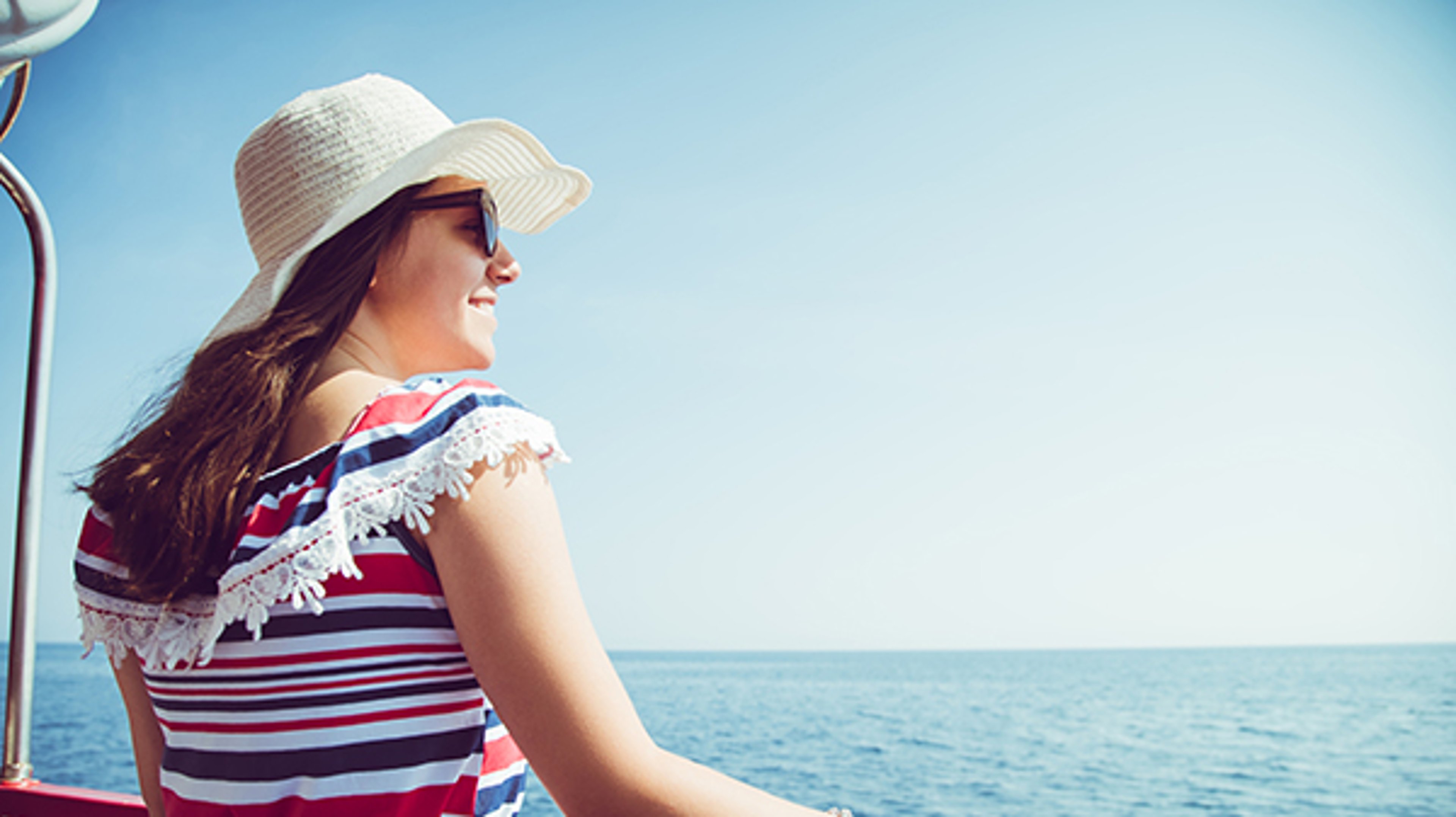 Woman smiling on a boat wearing a straw hat and sunglasses, looking at the ocean under a clear blue sky.