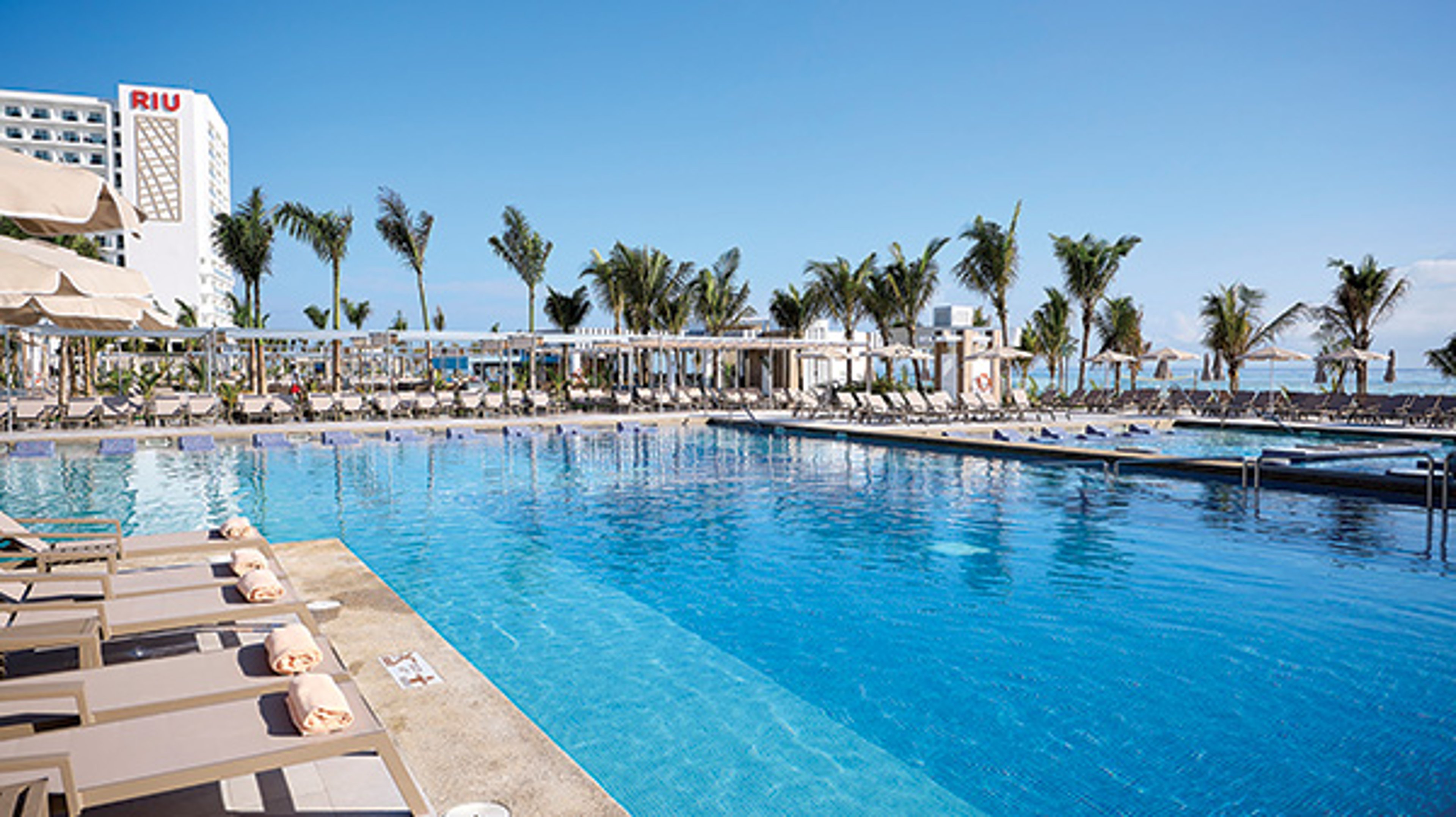 Large outdoor swimming pool at a resort with lounge chairs and a view of the ocean.