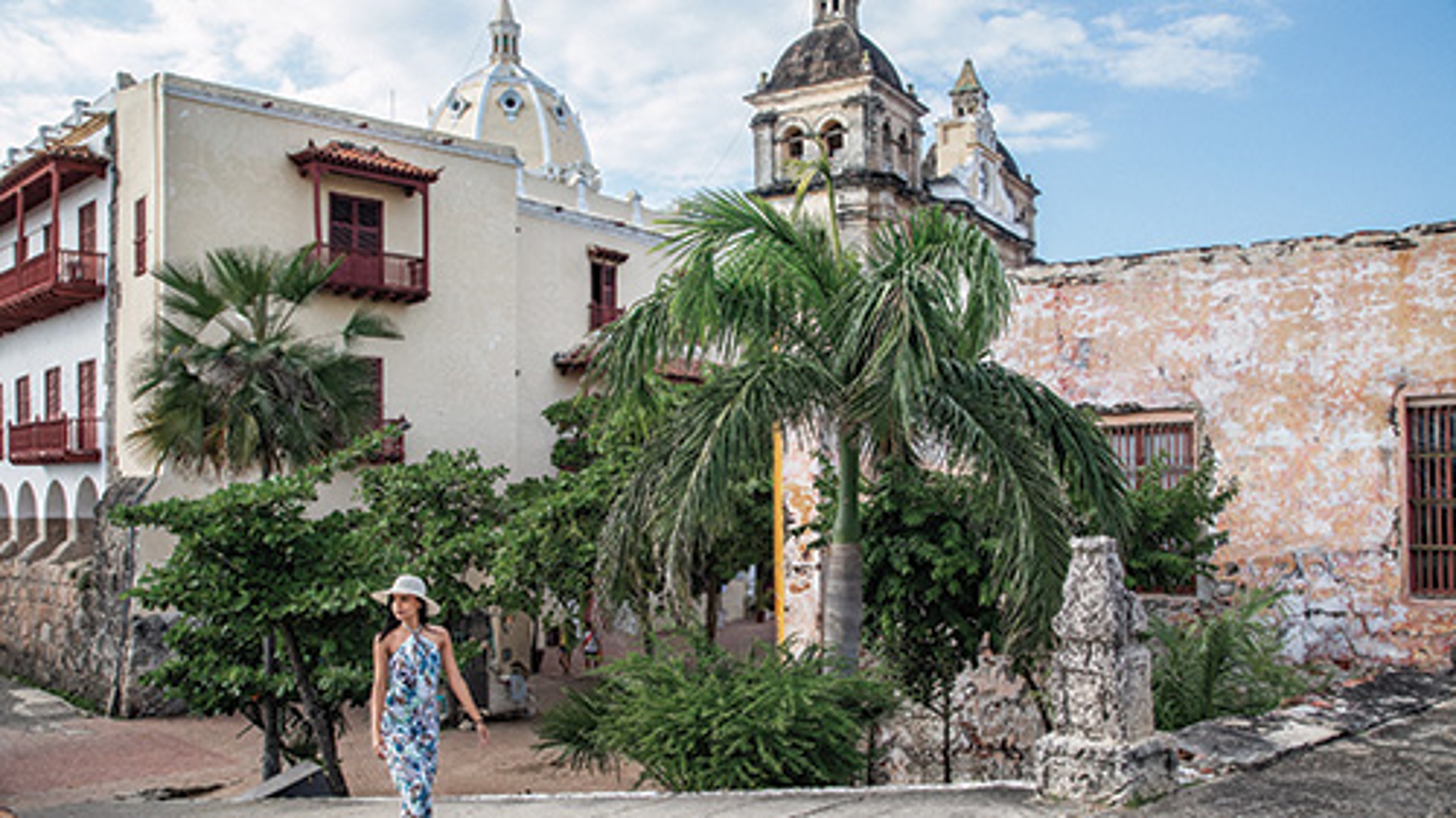 A woman walks in front of colonial-style buildings with red balconies and tropical greenery.