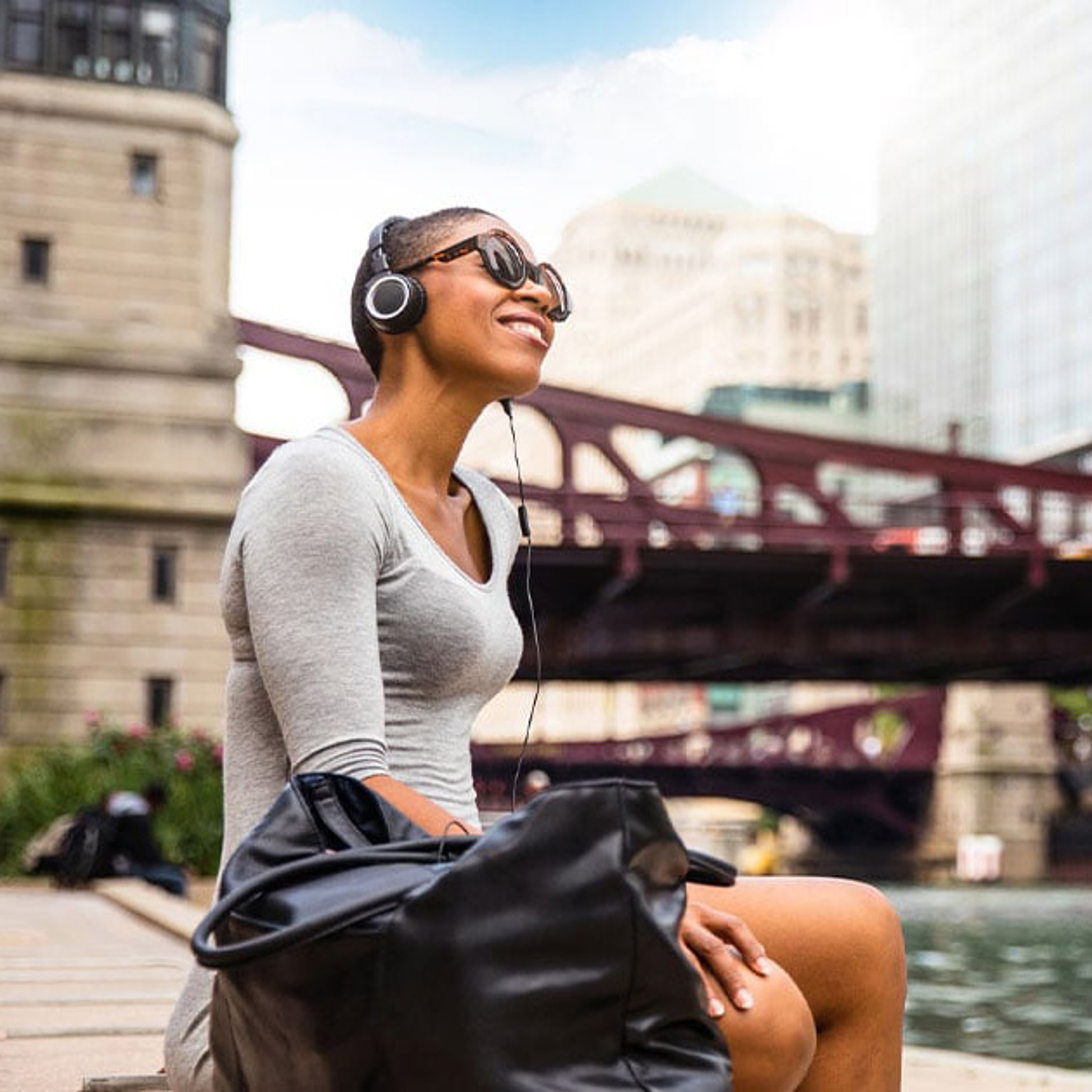 A smiling woman sits outdoors by a river, wearing headphones and sunglasses, with a black bag beside her.
