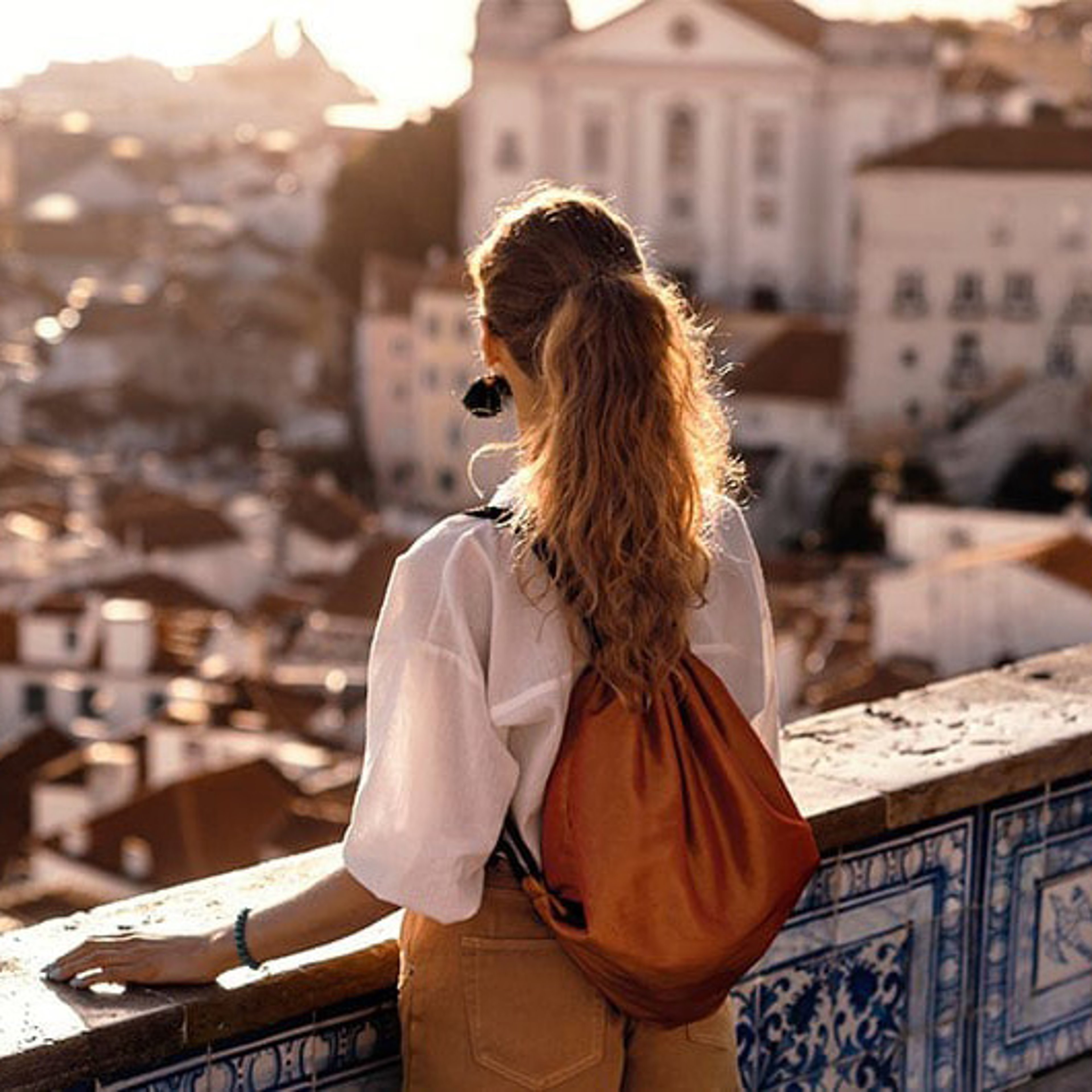 A woman viewed from behind looks out over a city landscape at sunset, leaning on a blue-tiled ledge.