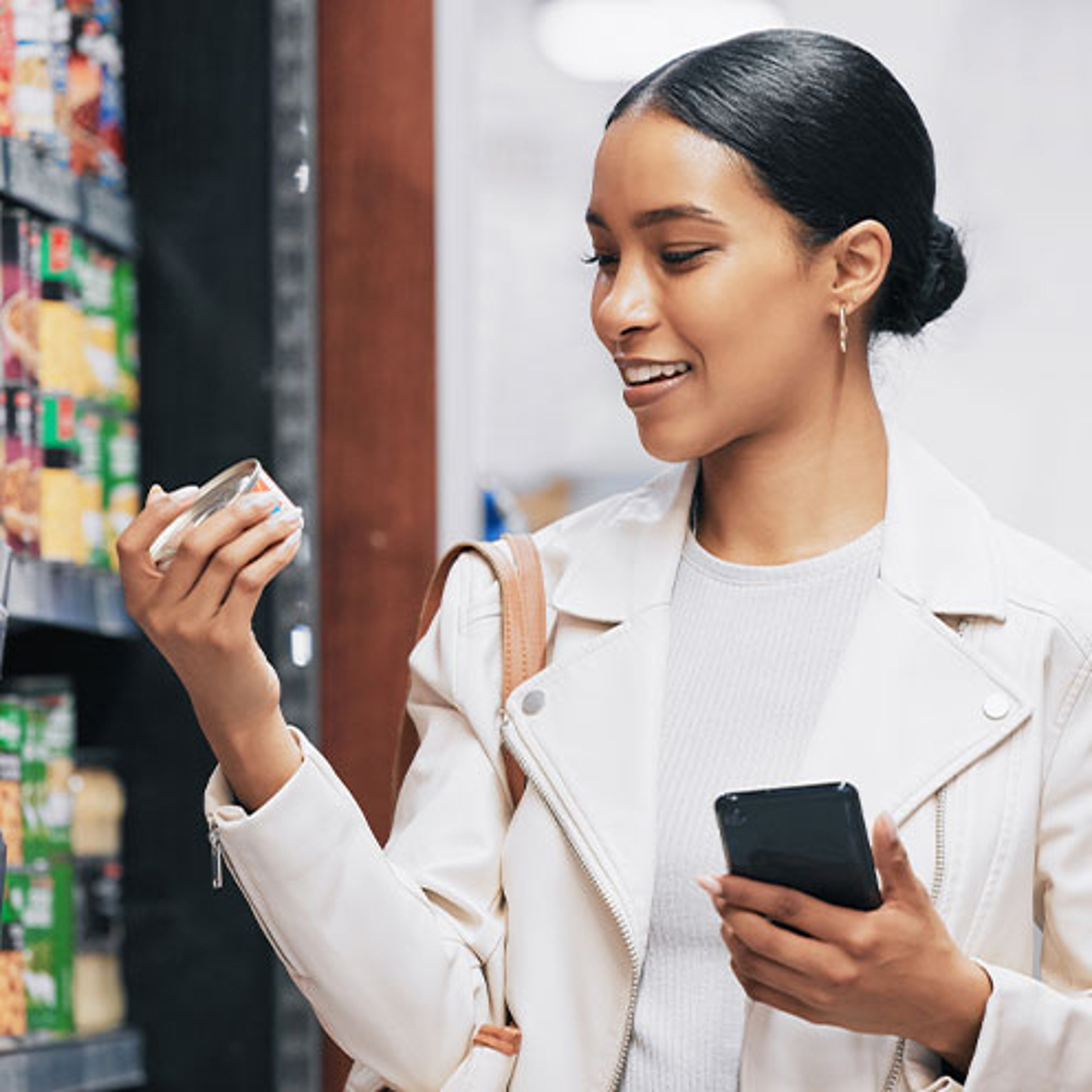 A smiling woman in a white jacket holds a small container and her smartphone while shopping in a grocery aisle.