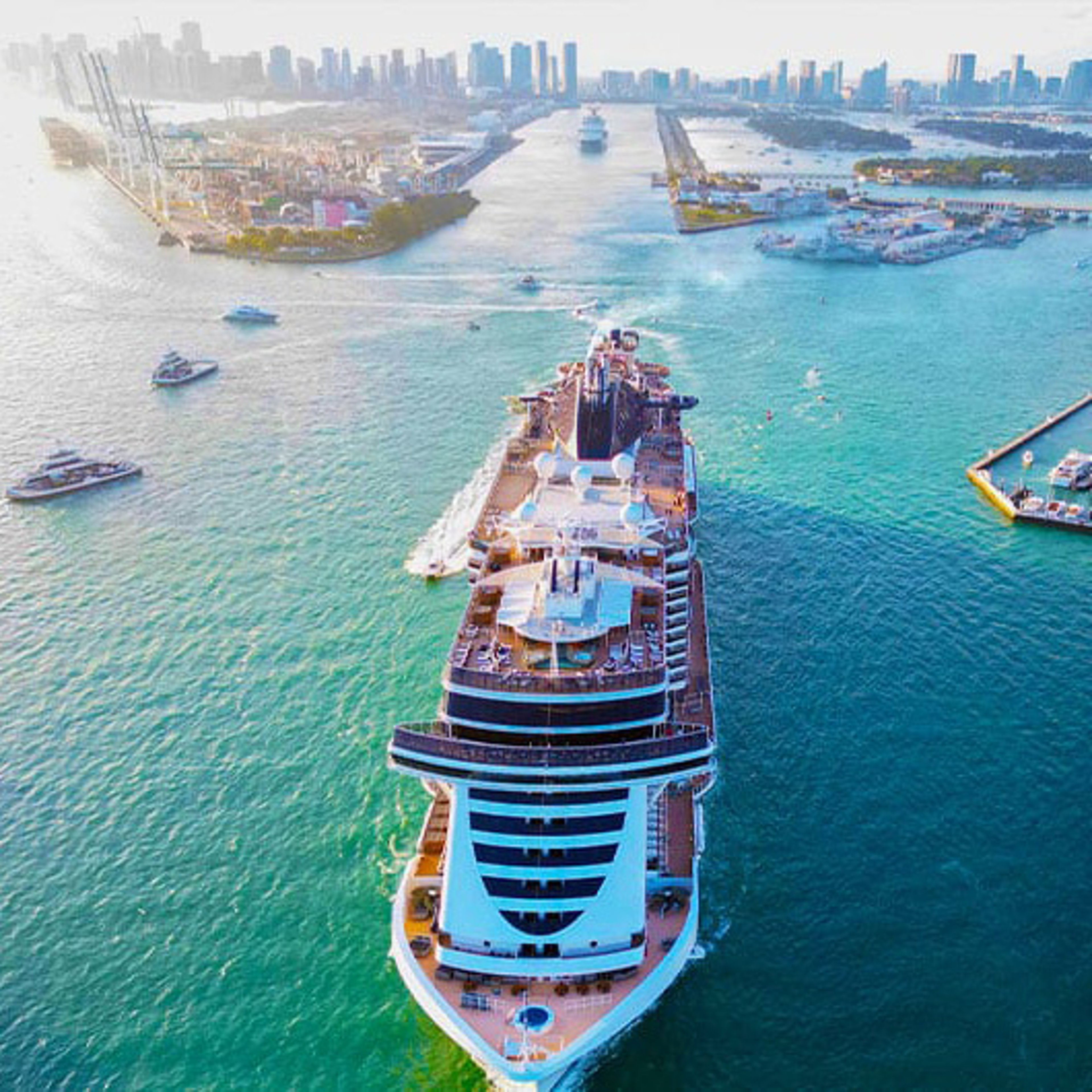 A large cruise ship departs a bustling port towards a city skyline in the distance, cutting through bright blue water.