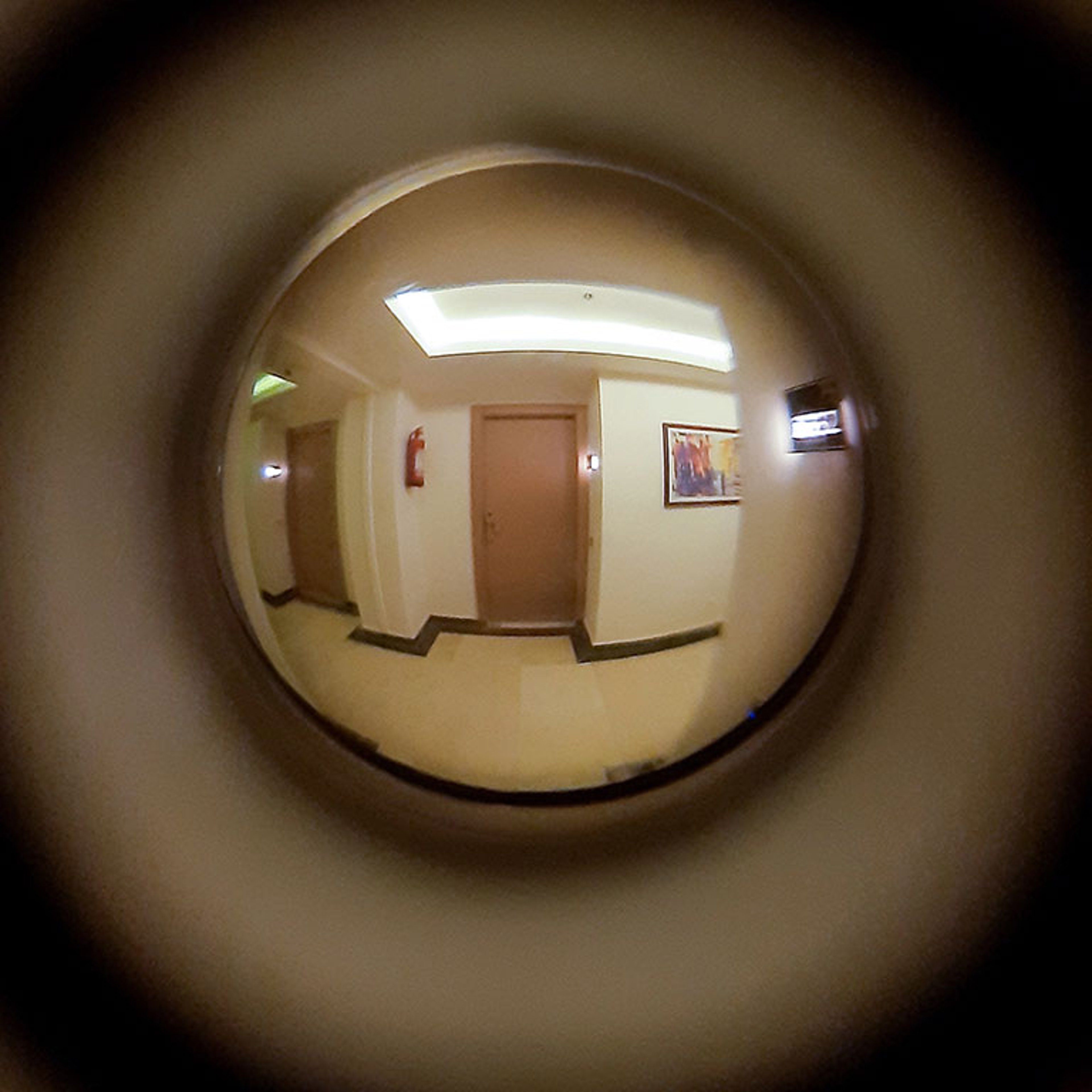  wide-angle, fish-eye view through a door peephole showing an empty hotel or apartment hallway with two doors and a fire extinguisher on the wall.