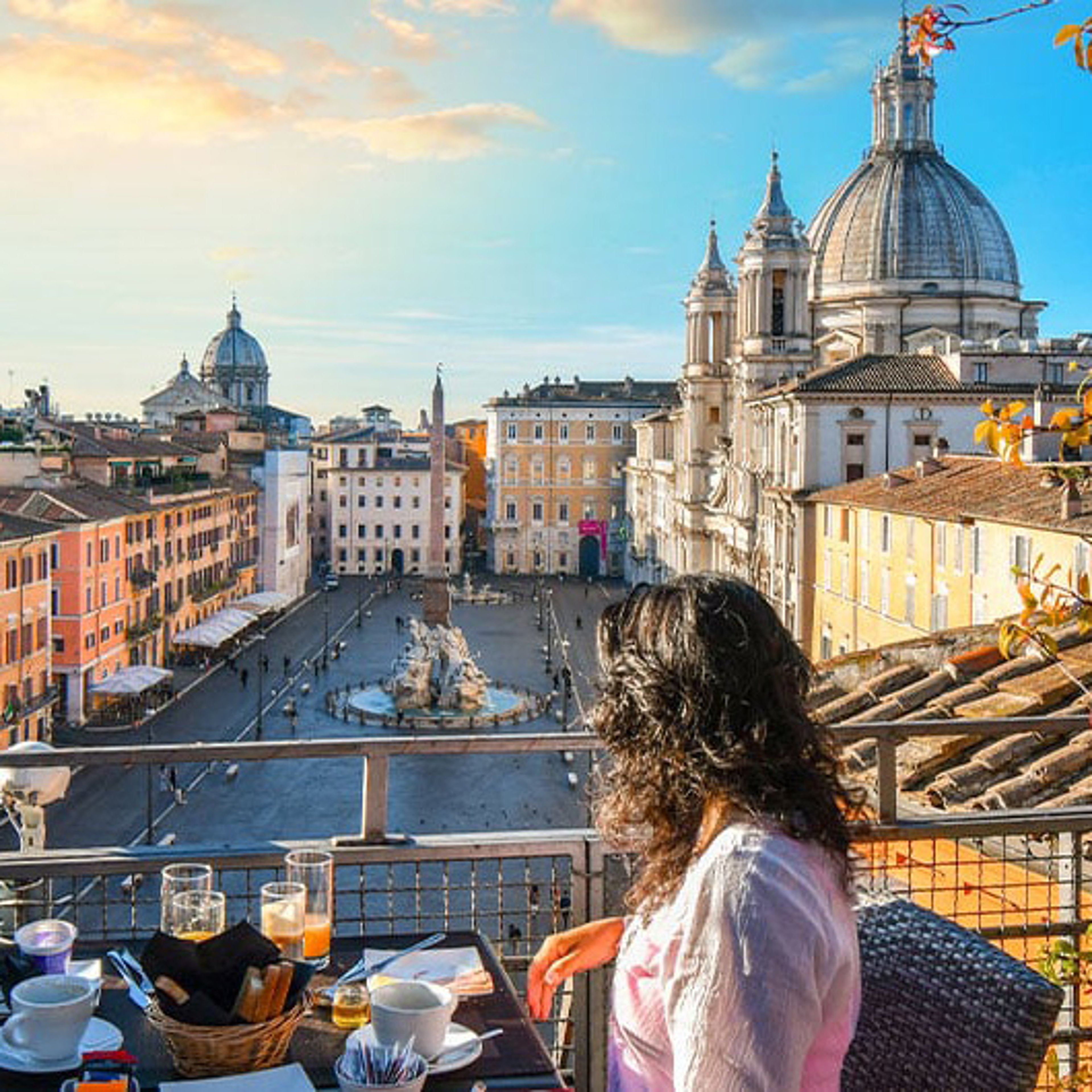 A woman viewed from behind sits at a breakfast table on a rooftop balcony, looking out over a historic city square with buildings and church domes.