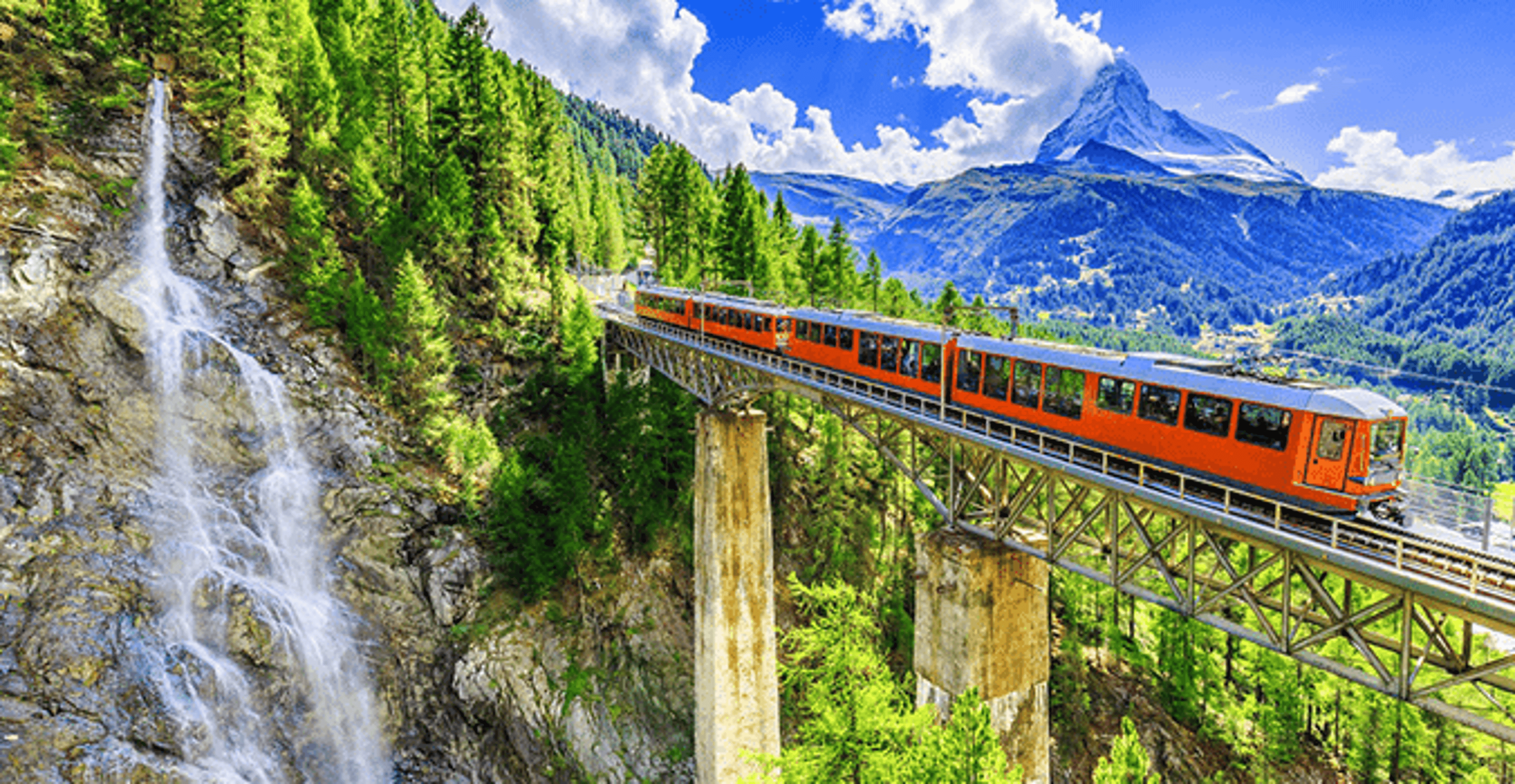 Train travelling across a rail bridge with mountains in the background and a waterfall on the cliffside