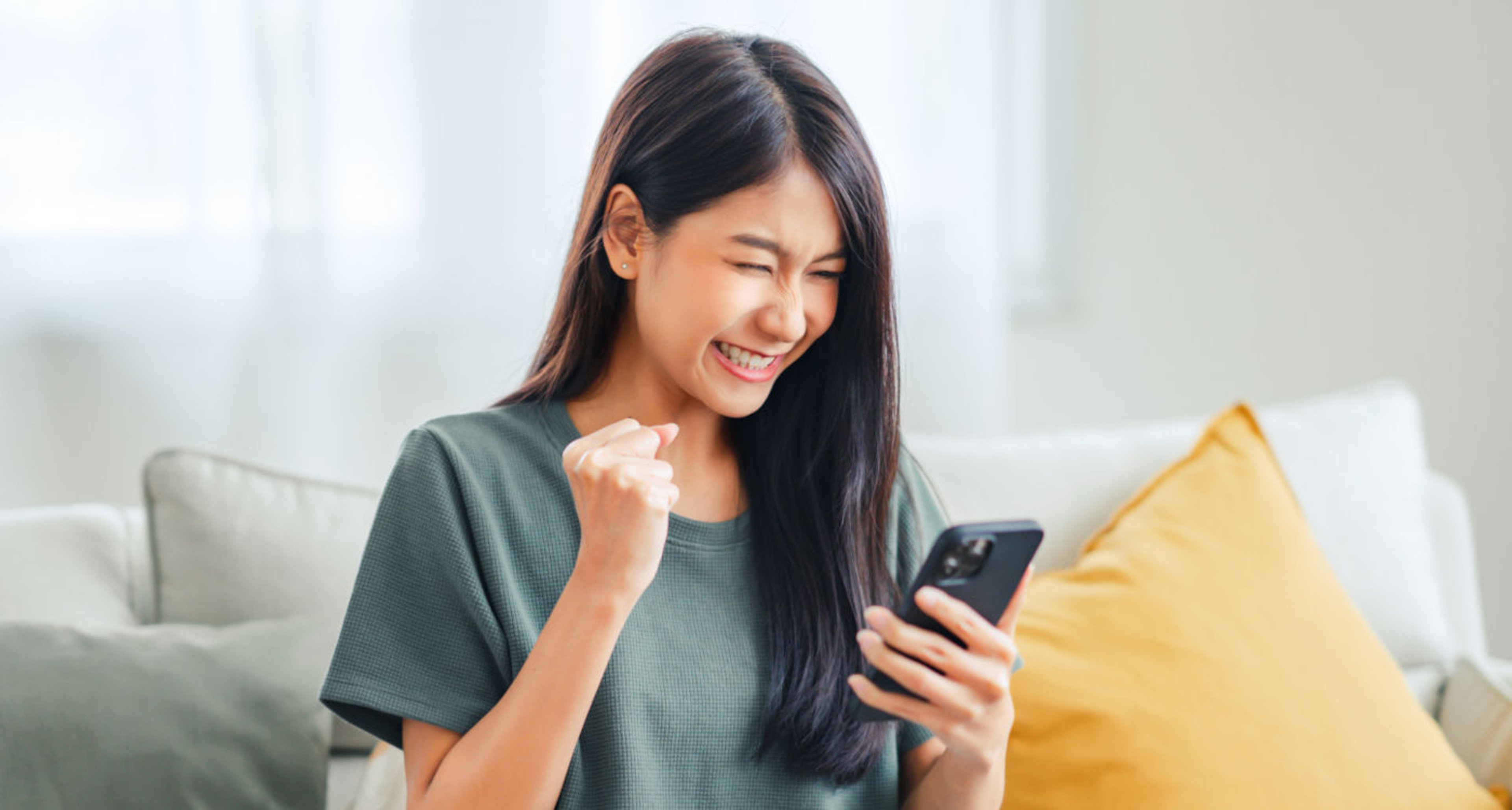 Woman sitting on couch smiling in celebration as she downloads mobile app on smartphone