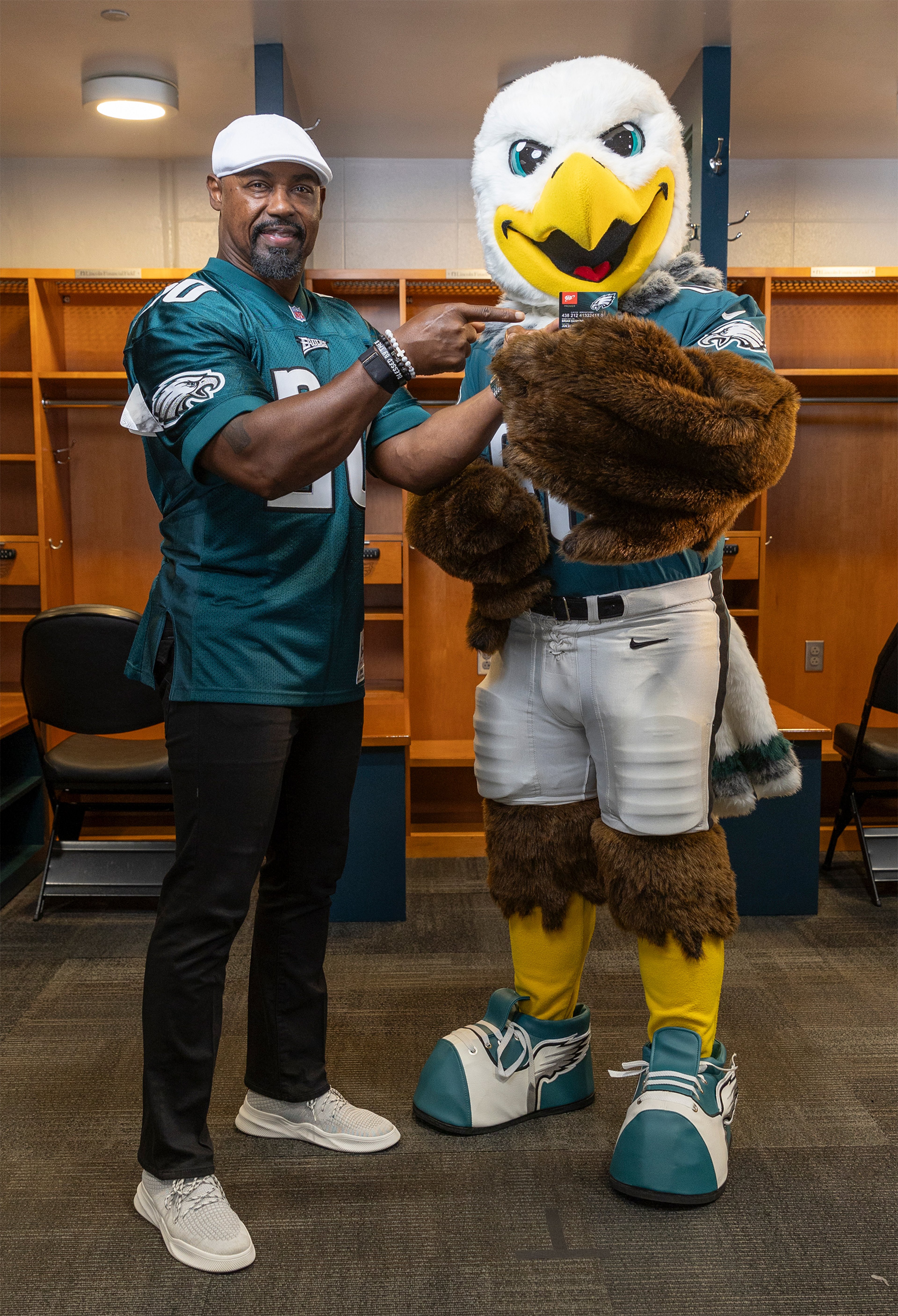 Philadelphia Eagles legend Brian Dawkins pointing at Eagles mascot Swoop holding a AAA Eagles MVP card while standing in Eagles locker room
