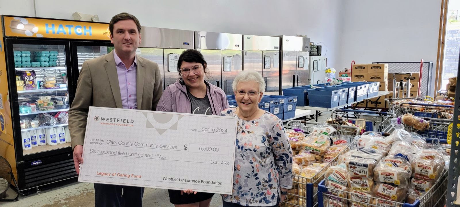 Three people holding a large check inside a food bank with groceries and crates visible in the background.