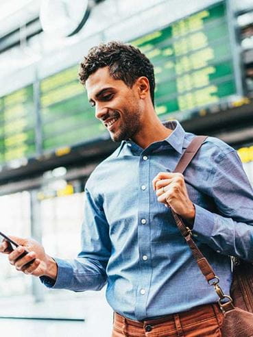 A smiling man checks his phone while carrying a shoulder bag at an airport terminal.