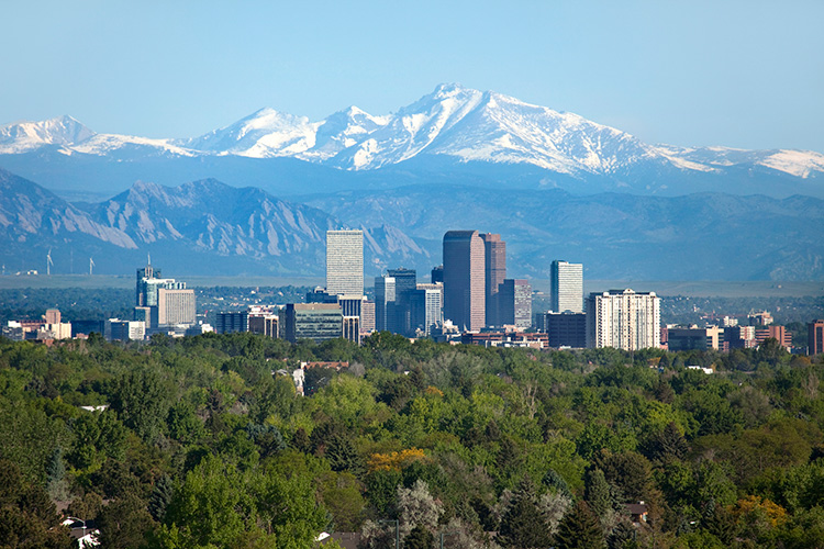 City skyline with skyscrapers in front of snow-capped mountains and lush greenery.