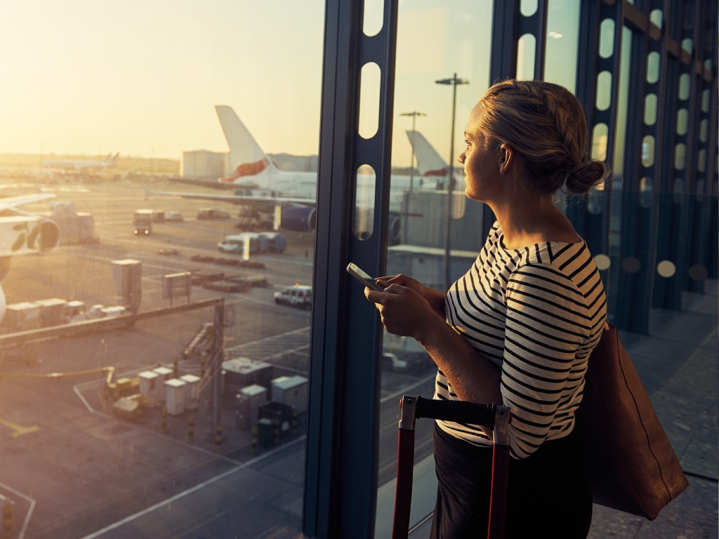 A woman holding her phone and luggage looks out an airport window at planes on the tarmac during sunset.