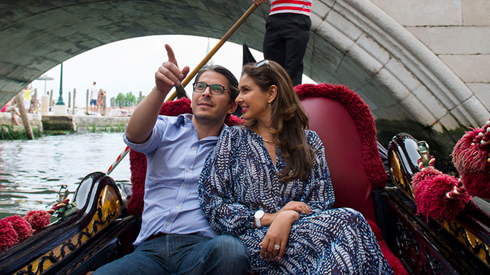 Couple sitting on a gondola under a stone bridge with a gondolier