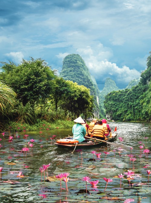 Tourists in a small boat navigating through a scenic river surrounded by lush greenery, mountains, and blooming pink water lilies under a partly cloudy sky.
