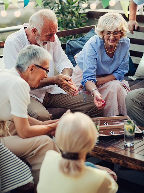 A diverse group of four older adults are gathered around a wooden table playing a board game, with expressions of joy and engagement.