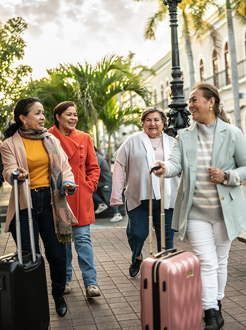 Four women walking together on a sidewalk, each with a suitcase. They are dressed in colorful, casual clothing with palm trees and a historic building in the background.