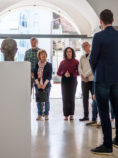 A group of five people in an art gallery, listening intently to a guide in front of a sculptural bust. Natural light fills the space through large windows.