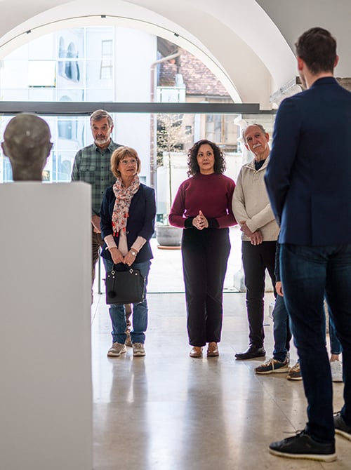 A group of five people in an art gallery, listening intently to a guide in front of a sculptural bust. Natural light fills the space through large windows.