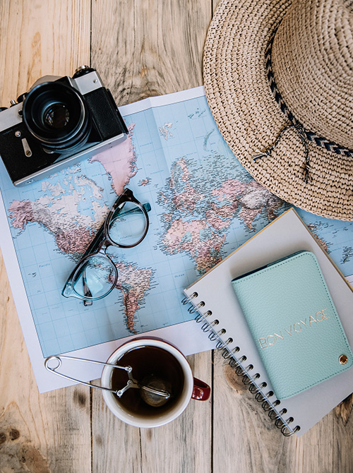 Flat lay of a map, camera, glasses, hat, notebook, passport holder, and a cup of tea.