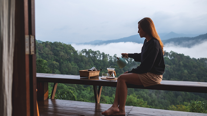 A young woman making drip coffee on a wooden balcony or deck, with a view of a lush green mountain landscape partially covered by mist and clouds.