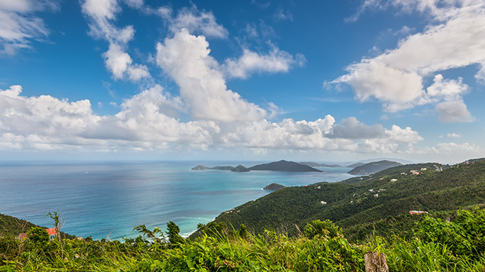  A high-angle scenic view from a green, grassy hillside overlooking multiple lush tropical islands and the blue ocean under a bright sky with white clouds.