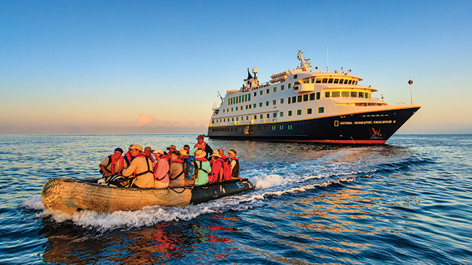 A small inflatable boat full of tourists wearing life vests cruising on the ocean away from the large expedition ship National Geographic Endeavour II under a sunset or sunrise sky.