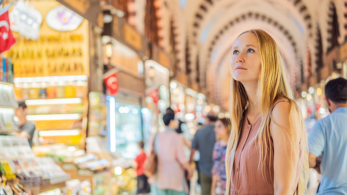 A woman with long blonde hair standing inside a large, ornate indoor market with arched ceilings, looking up, surrounded by various stalls and blurred figures of other people.
