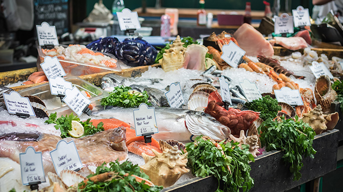 A vibrant market stall display piled high with various fresh seafood on ice, including whole fish, lobsters, scallops, and price tags.