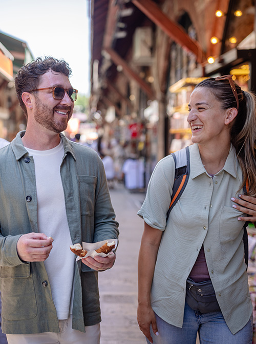 A man and a woman smiling and walking in a bustling outdoor market or alleyway, with the man holding a pastry or food item.