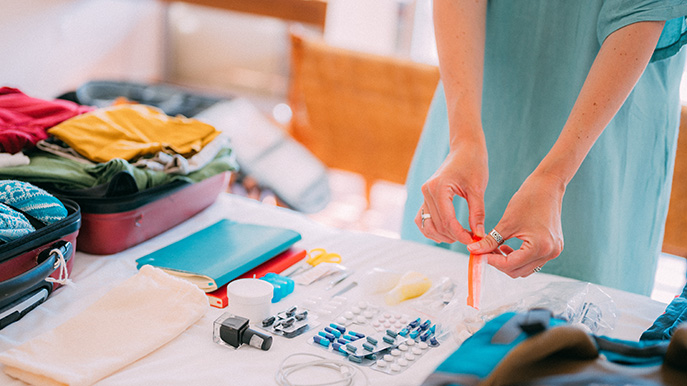 A person organizing items on a table with a suitcase, clothes, pills, and travel essentials