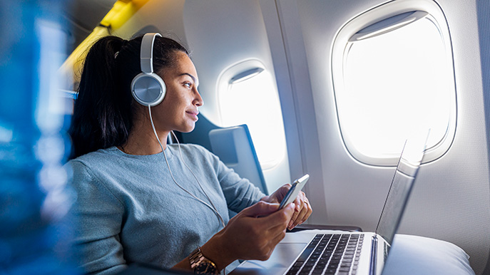 Woman wearing headphones, holding a smartphone, and using a laptop in an airplane seat by the window