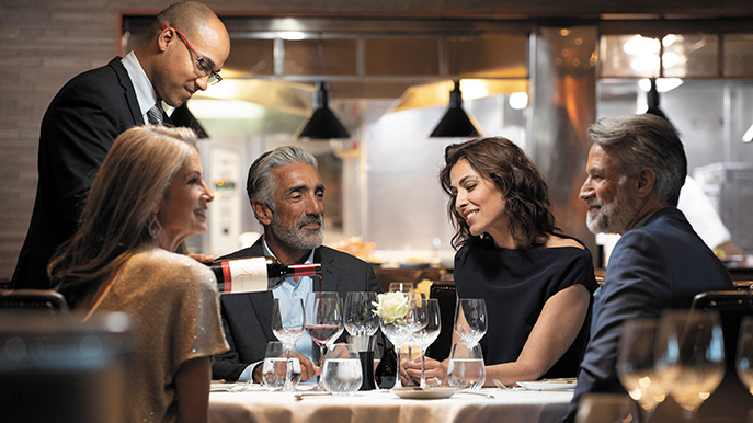 A group of four people dressed elegantly seated at a restaurant table with a server pouring wine