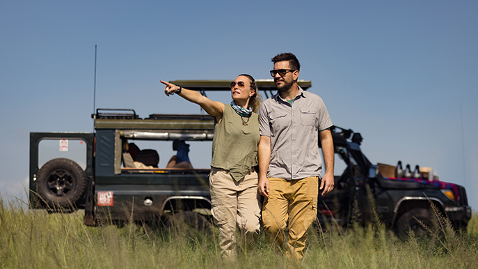 Couple stands outside open-top safari vehicle overlooking grassy savanna