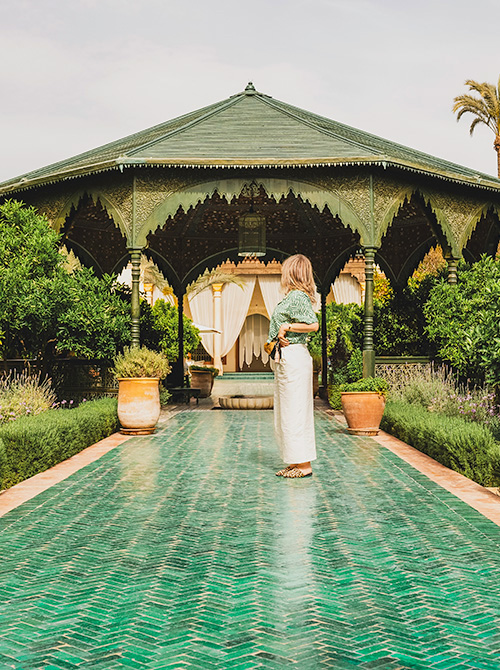 Woman standing on a patterned green tile path in Le Jardin Secret, Marrakech, with a traditional Moroccan pavilion and lush greenery in the background