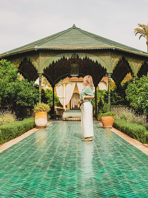 Woman standing on a patterned green tile path in Le Jardin Secret, Marrakech, with a traditional Moroccan pavilion and lush greenery in the background