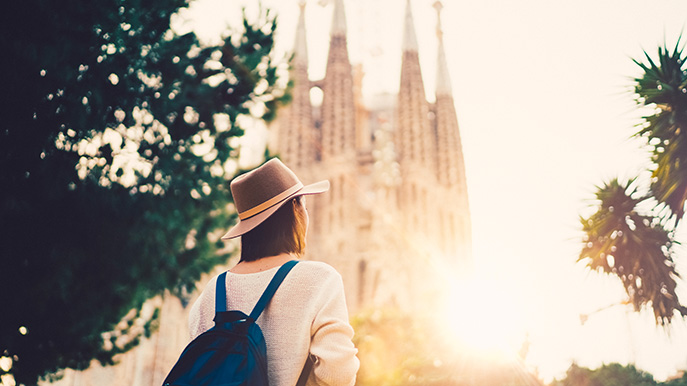 A person wearing a hat and backpack stands with their back to the camera, looking at the towering spires of the Sagrada Familia basilica in Barcelona, under a bright, sunny sky