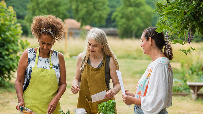 Three women in aprons are gathered around a table outdoors, surrounded by fresh produce and cooking ingredients, appearing to be preparing a meal or discussing a recipe