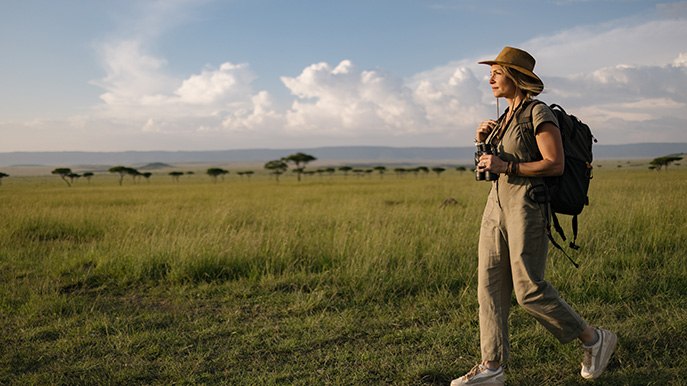 A woman in a safari hat and outfit, with a backpack and binoculars, walks through the grassy plains of an African savanna