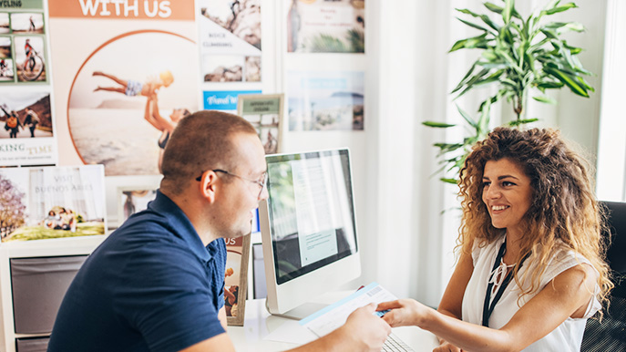 Travel advisor handing a brochure to a client in an office setting with travel posters on the walls
