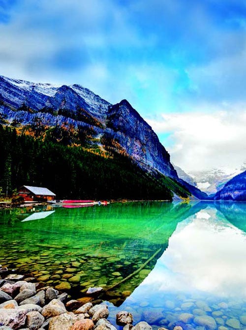 Snow-dusted mountains and evergreen forest reflected in a clear turquoise alpine lake, with a small lakeside cabin and boats near the rocky shore.