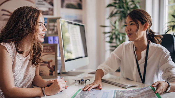 Two women at a desk discussing an open brochure with a model airplane and computer nearby
