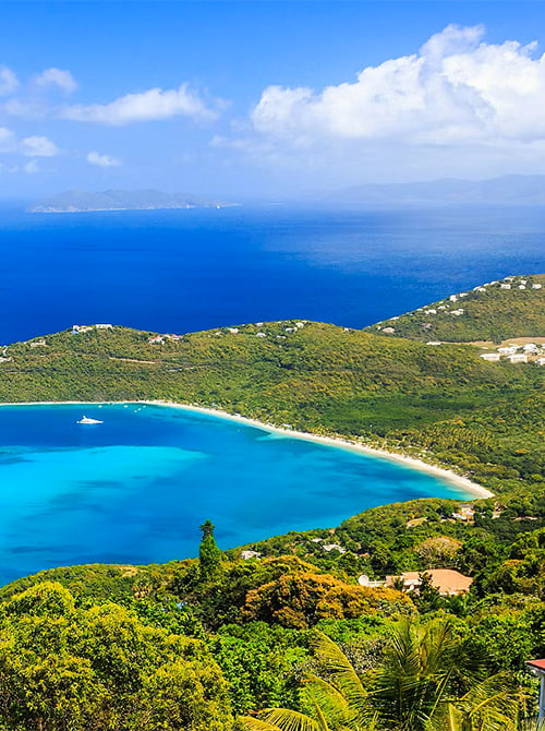 Turquoise bay with a curved sandy beach, lush green hills and sailboats on the water.